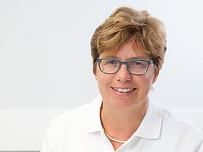 Freundliche Ärztin in weißer Arbeitskleidung sitzt an einem Schreibtisch in einer Praxis, lächelt; vor ihr liegen ein offenes Buch und ein Stethoskop. Die Szene vermittelt Kompetenz und vertrauensvolle medizinische Beratung.