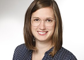 Sabrina Maier Porträt einer lächelnden Person mit runder Brille und schulterlangem braunem Haar. Die Person trägt ein dunkelblaues, weiß gepunktetes Hemd vor hellem, neutralem Hintergrund.