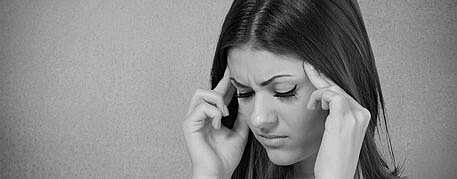 Schwarzweißfoto: Frau hält sich mit beiden Händen die Schläfen, Augen geschlossen, schmerzhaftes Gesicht. Deutet auf Kopfschmerzen oder Stress.