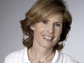 Porträt einer lächelnden Frau mit hellbraunen, leicht welligen Haaren, weißem Poloshirt und Perlenkette vor neutral grauem Hintergrund.