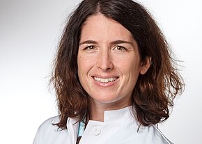 Porträt einer lächelnden Frau in weißem Kittel vor hellem Hintergrund. Mittellange, braune, leicht wellige Haare; freundlicher Blick. Wirkt wie medizinisches Fachpersonal.