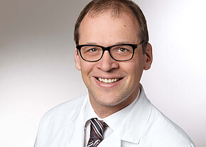 Porträt eines lächelnden Mannes mit Brille in weißem Arztkittel und gestreifter Krawatte vor neutralem Hintergrund.