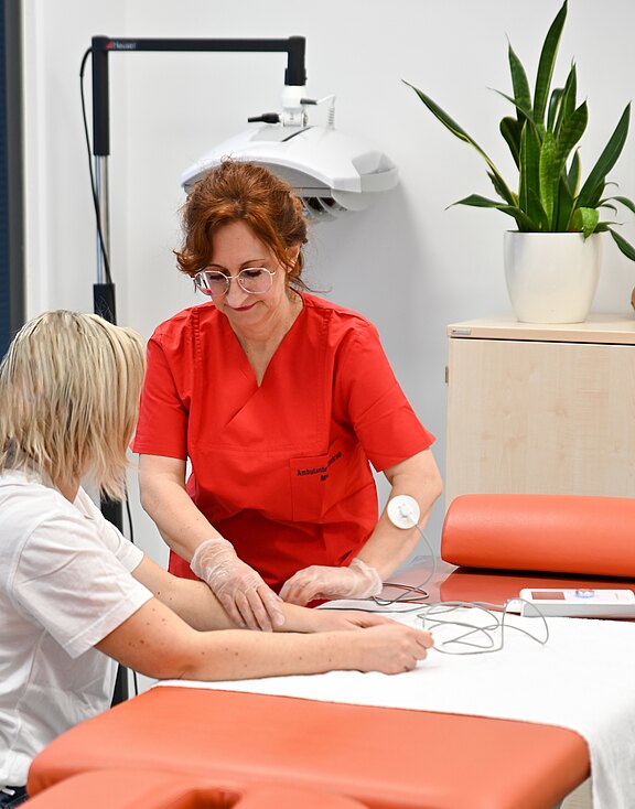 Eine medizinische Fachkraft mit Handschuhen in roter Arbeitskleidung untersucht den Arm einer sitzenden Patientin und schließt Elektroden oder Kabel an. Beide befinden sich in einem Behandlungsraum auf einer Untersuchungsliege. Fokus auf Betreuung und Diagnostik.