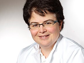 Porträt einer lächelnden Person mit kurzen dunklen Haaren und Brille, in einem weißen Kittel, vor neutralem Hintergrund. Halbnaher Kopf-Schulter-Ausschnitt mit professioneller Erscheinung.