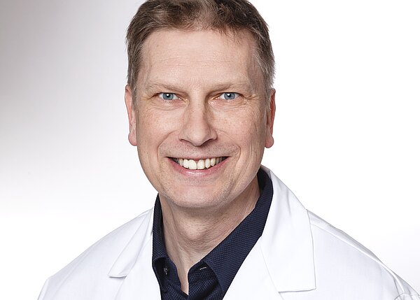 Porträt eines lächelnden Mannes mittleren Alters in weißem Kittel vor hellem Hintergrund; Kopf-und-Schulteraufnahme, direkter Blick in die Kamera, freundlicher, professioneller Eindruck.