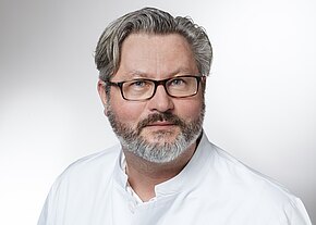 Dr. med. Maximilian Keil Porträt eines Mannes mit Brille und grauem Haar, kurzer Vollbart. Er trägt einen weißen Kittel und blickt ruhig in die Kamera vor neutralem Hintergrund.