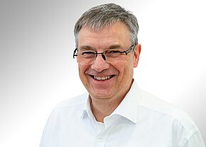 Porträt eines lächelnden Mannes mittleren Alters mit kurzem grauem Haar und Brille, in weißem Hemd, vor hellem neutralem Hintergrund, freundlich in die Kamera blickend.