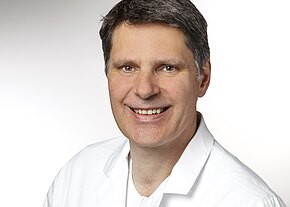 Porträtfoto eines mittelalten Mannes mit kurzen dunklen Haaren, der freundlich lächelt. Er trägt einen weißen Kittel vor neutral hellem Hintergrund.