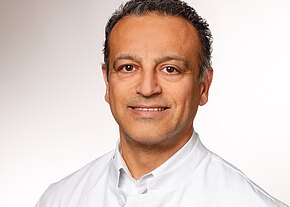 Shafie Ariai Ein lächelnder Mann trägt einen weißen Kittel. Sein Gesicht ist freundlich und er hat dunkle, leicht wellige Haare. Der Hintergrund ist schlicht und hell. Die Darstellung vermittelt einen professionellen Eindruck, der häufig mit medizinischen Berufen assoziiert wird.