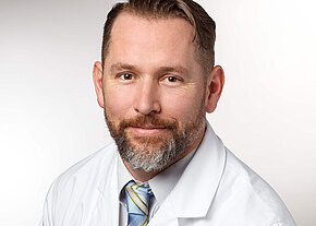 Porträt eines erwachsenen Mannes mit kurzem Haar und Bart, leichtes Lächeln. Er trägt weißen Arztkittel, Hemd und gestreifte Krawatte vor neutralem Hintergrund.