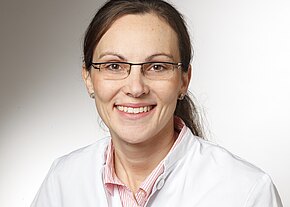 Porträt einer lächelnden Frau mit Brille in weißem Kittel vor neutralem Hintergrund; freundlicher Blick, leicht nach rechts gedreht.