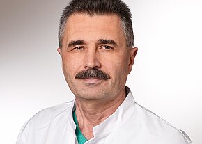 Ein männlicher Arzt mit Schnurrbart trägt einen weißen Kittel und ein grünes Oberteil. Er blickt freundlich in die Kamera. Der Hintergrund ist neutral und hell, was eine professionelle Atmosphäre vermittelt.