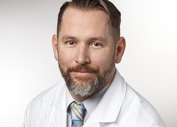 Porträt eines Mannes mittleren Alters mit kurzem Haar und Bart, der einen weißen Kittel und eine Krawatte trägt. Er blickt freundlich in die Kamera vor neutralem Hintergrund.