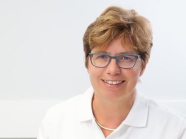 Freundliche Ärztin in weißer Arbeitskleidung sitzt an einem Schreibtisch in einer Praxis, lächelt; vor ihr liegen ein offenes Buch und ein Stethoskop. Die Szene vermittelt Kompetenz und vertrauensvolle medizinische Beratung.