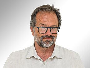 Dr. Klaus Pfaffenberger Ein Mann mit einem kurzen, grauen Bart und einer Brille blickt direkt in die Kamera. Er trägt ein helles, khakifarbenes Hemd. Der Hintergrund ist hell und verschwommen, was den Fokus auf sein Gesicht lenkt.