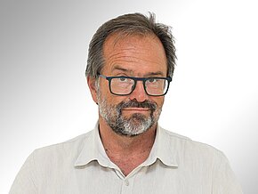 Ein Mann mit einem kurzen, grauen Bart und einer Brille blickt direkt in die Kamera. Er trägt ein helles, khakifarbenes Hemd. Der Hintergrund ist hell und verschwommen, was den Fokus auf sein Gesicht lenkt.