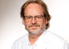 Dr. med. Winfrid Melzner Frontales Porträt eines mittelalten Mannes mit Brille, kurzen welligen Haaren und grauem Bart. Er trägt einen weißen Kittel und Hemd und blickt ruhig in die Kamera vor hellem, neutralem Hintergrund.