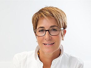 Ärztin im weißen Kittel sitzt lächelnd an einem Schreibtisch, hält einen Stift und schreibt auf ein Formular. Neben ihr liegen Stethoskop und Tastatur in einer hellen Praxis.